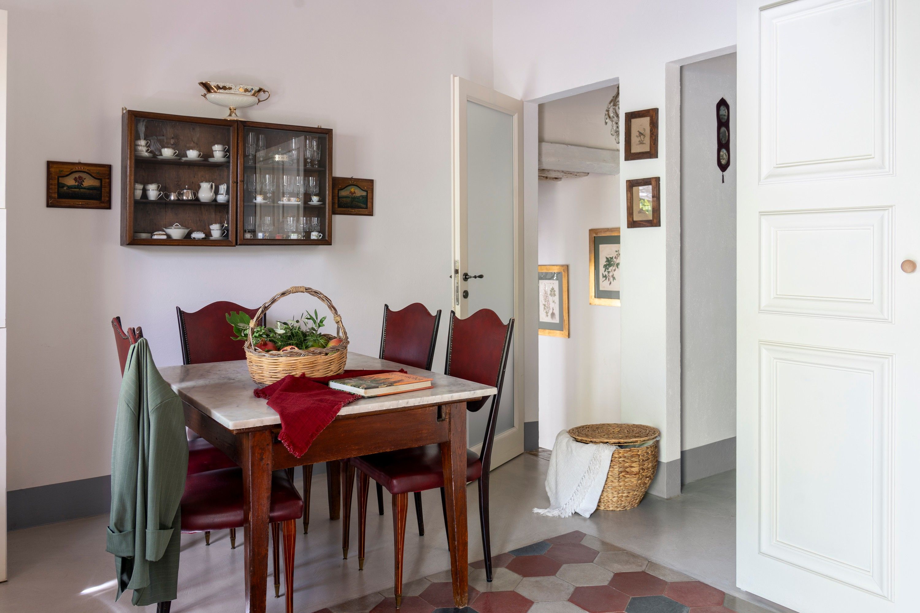Un comedor con una mesa de madera, sillas tapizadas en rojo, una cesta de mimbre con frutas y un libro.