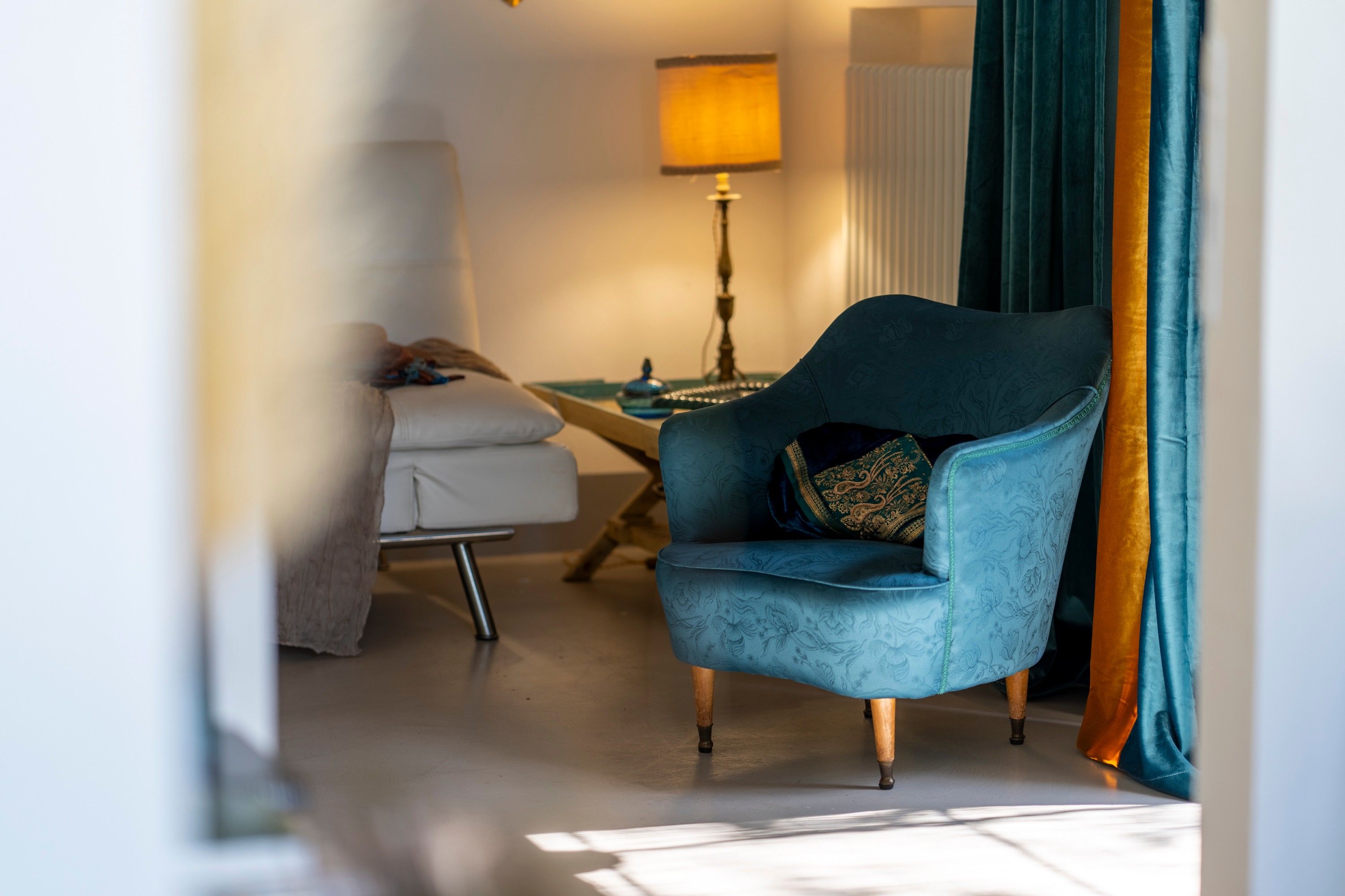 A room with a blue armchair, a white sofa, a lamp, and a window with curtains.