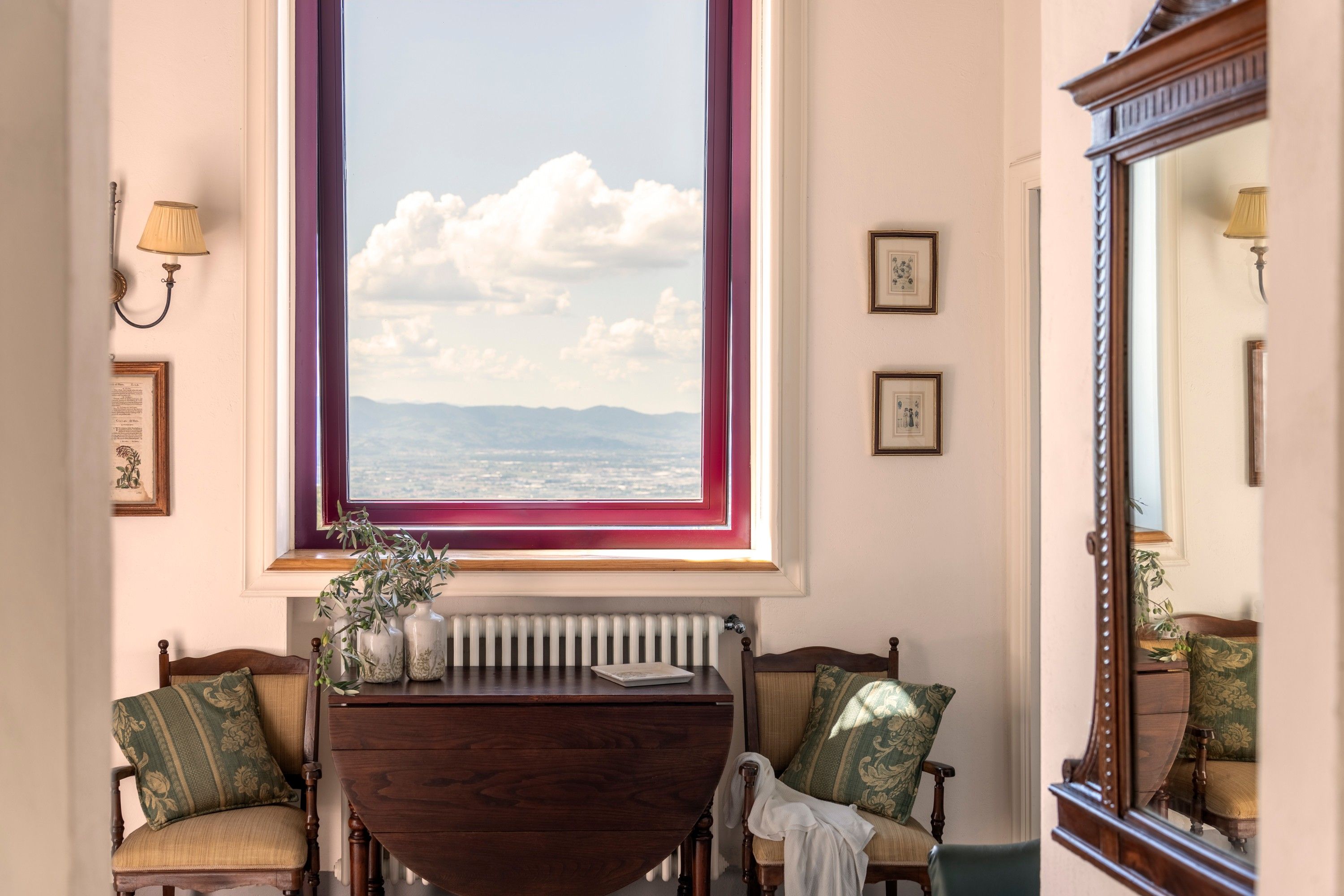 A room with a large window, a wooden table, two chairs, a radiator, and framed pictures on the wall.