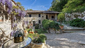 Una casa in pietra con giardino, un vialetto sterrato e un tavolo in vetro con un vaso di fiori.