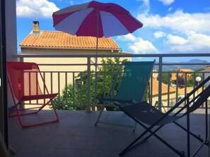 Ein Balkon mit zwei Stühlen, einem rot-weißen Sonnenschirm und Blick auf Gebäude und Bäume.