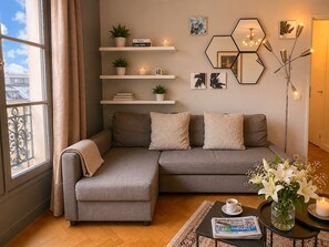 Uma sala de estar moderna com um sofá seccional cinza, uma mesa de centro, um vaso com flores, um abajur e uma estante de livros com livros e plantas.