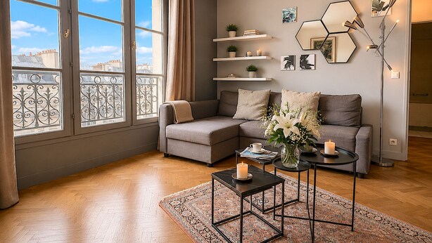 Una sala de estar moderna con un sofá gris, una mesa de centro y una lámpara de pie. Hay estanterías con objetos decorativos y vistas de la ciudad a través de amplios ventanales.