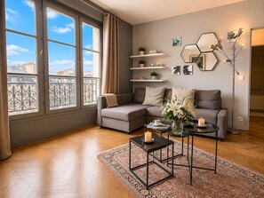 Ein modernes Wohnzimmer mit grauem Sofa, Couchtisch und Stehlampe. Regale mit Dekoration und Blick auf die Stadt durch große Fenster.