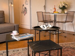 Ein Wohnzimmer mit einer Couch, einem Couchtisch mit Zeitungen, einem kleinen Tisch mit einer Kerze sowie einem Essbereich mit Stühlen und einem Tisch, gedeckt mit einer Flasche, Gläsern und Blumen.