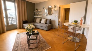 Una sala de estar con un sofá, una mesa de centro con flores, un comedor con mesa y sillas, y un gran ventanal con cortinas.