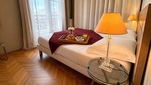 Habitación de hotel con cama, mesita de vidrio con lámpara, bandeja con croissants y café, y ventana con cortinas.