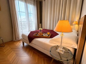 Ein Hotelzimmer mit einem Bett, einem Nachttisch mit Lampe, einer Ablage mit Croissants und Kaffee sowie einem Fenster mit Vorhängen.