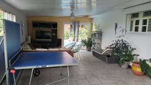 Ein geräumiger Wohnbereich mit Tischtennisplatte, einer Couch und einem Fernseher. Üchtischpflanzen und ein Bücherregal sind ebenfalls vorhanden.