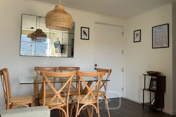A dining area with a glass table and wooden chairs, a woven pendant light, and framed pictures on the walls.