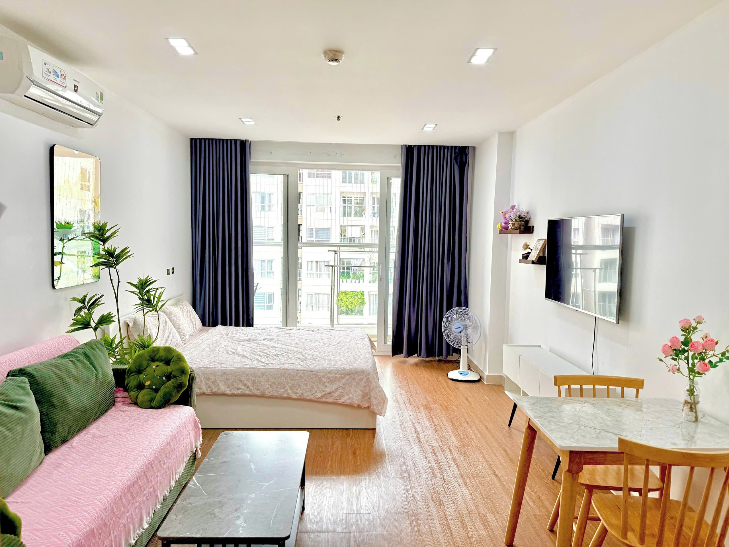 A hotel room with a bed, a sofa, a small table, a fan, a TV, and a balcony with curtains.
