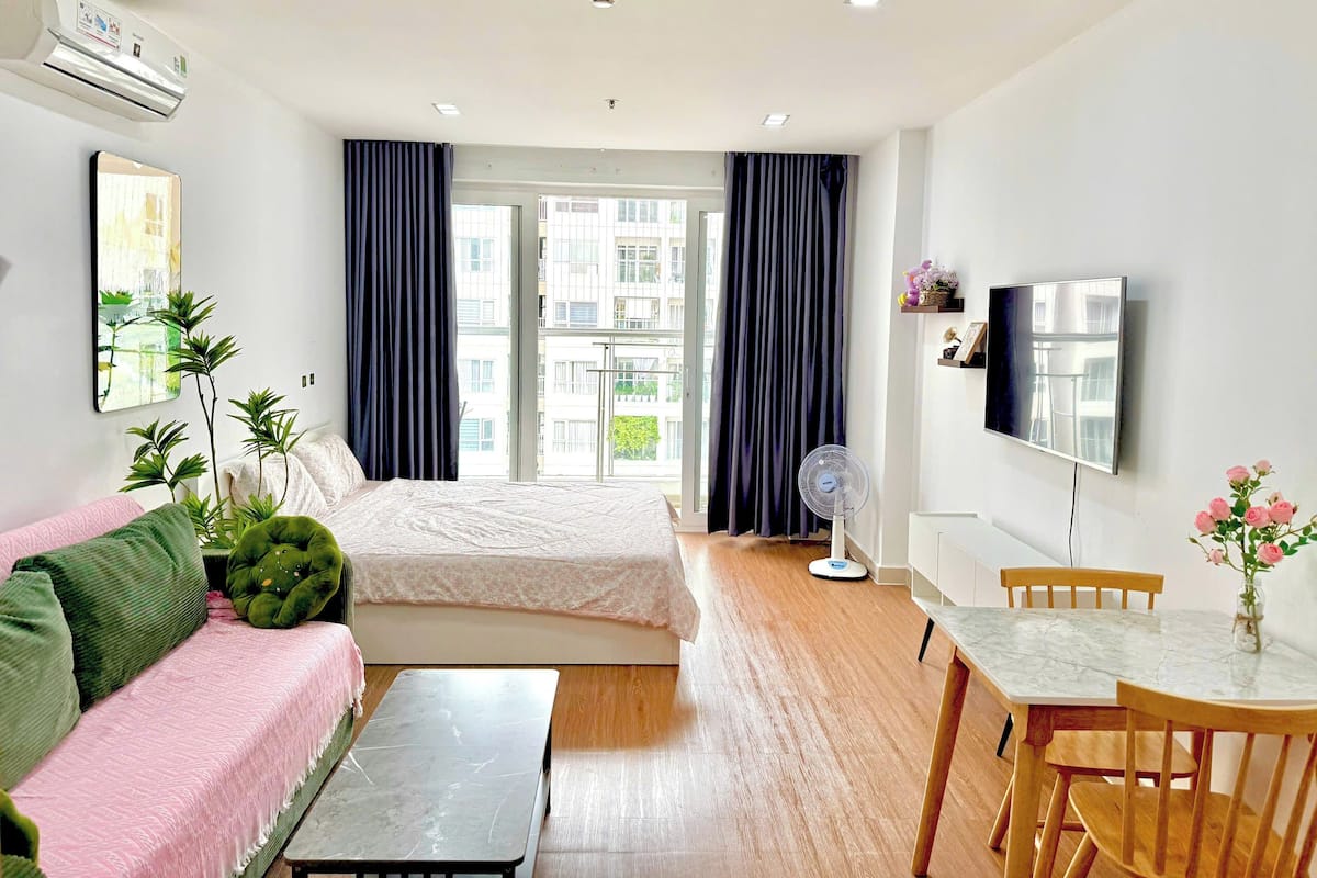A hotel room with a bed, a sofa, a small table, a fan, a TV, and a balcony with curtains.