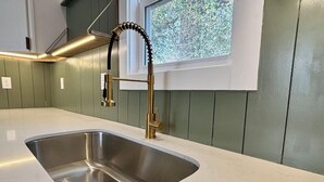 A kitchen with a stainless steel sink, a gold faucet, and a window with a view of greenery. - Brooklyn Arts District Historic Home 1BR, 1BA (Wilmington)