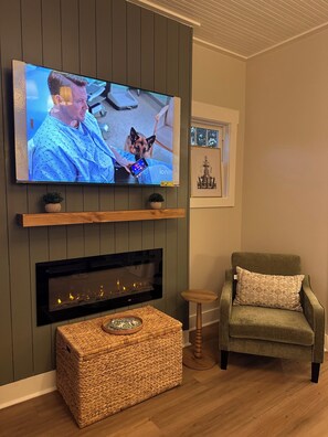 Ett vardagsrum med en platt-TV som hänger på väggen, en öppen spis under, en grön fåtölj och ett litet bord med en korg.