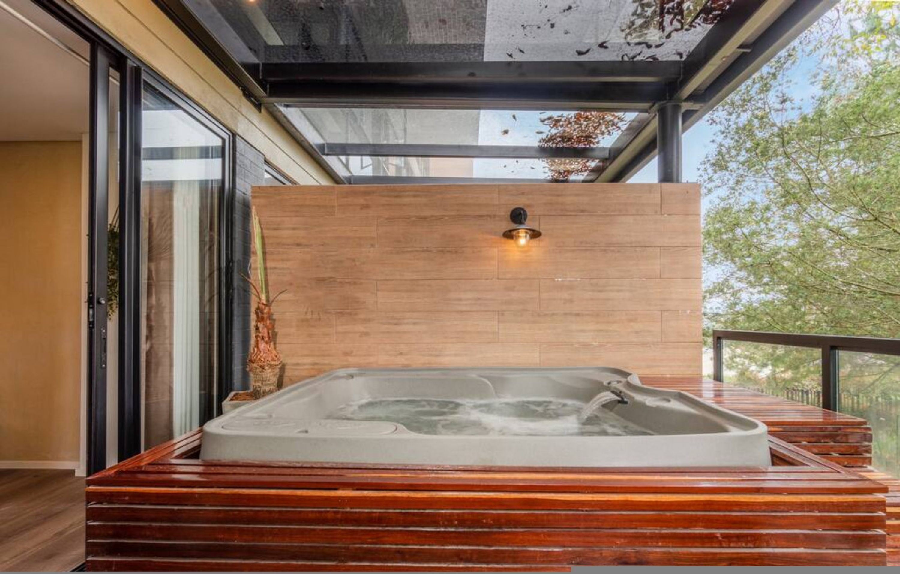 Un jacuzzi con plataforma de madera, una luz empotrada en la pared y una puerta de cristal.