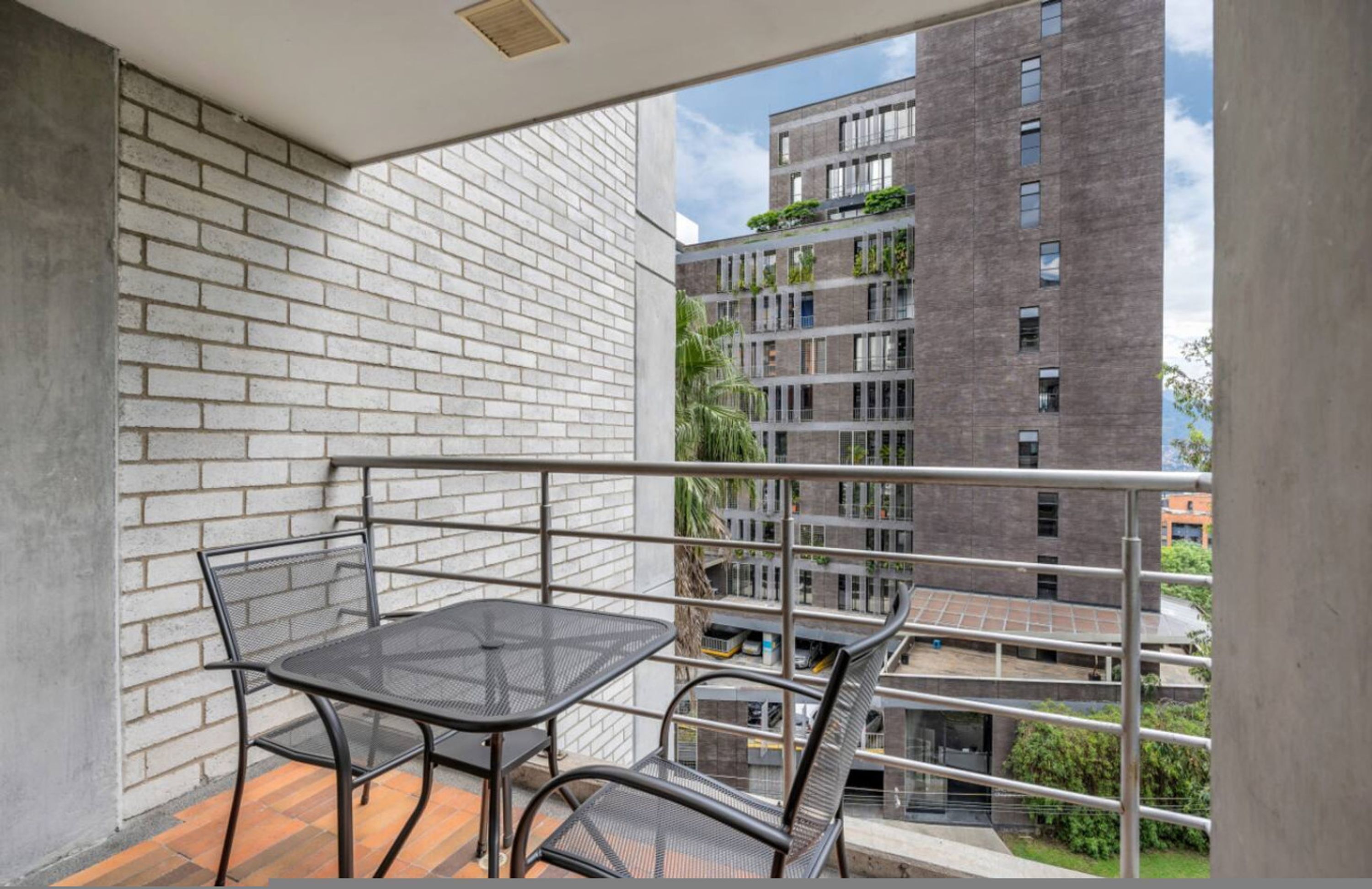 Ein Balkon mit einem Metalltisch und -stühlen, einer weißen Ziegelwand und Blick auf ein mehrstöckiges Gebäude mit Grünflächen.