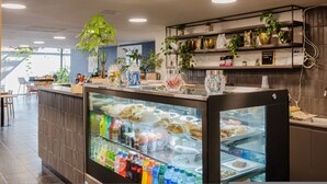 A modern cafe interior with a glass display case of pastries, a counter with a variety of drinks, and shelves with plants and kitchen items. - Cyan 703 - 2BR in Medell n (Medellín)