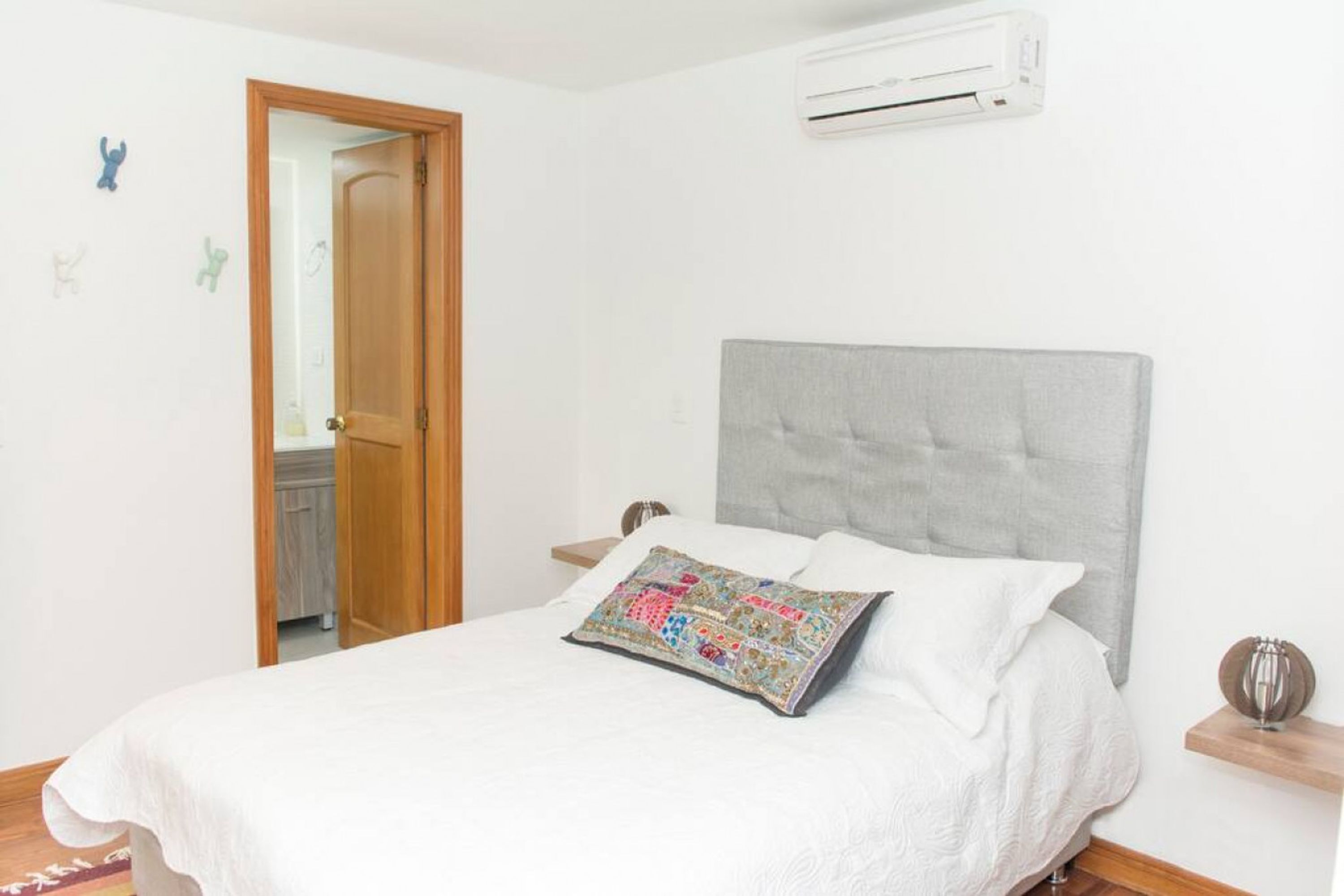 A bedroom with a bed, a grey upholstered headboard, a white bedspread, a colorful pillow, a wooden nightstand, and a wall-mounted air conditioner.