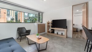 Un salon moderne avec un canapé, une table basse, un téléviseur posé sur un meuble en bois et une vue sur la ville à travers de grandes fenêtres.
