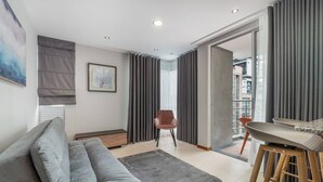 A modern living room with a grey sofa, a wooden coffee table, and a chair. There is a large window with curtains, a painting on the wall, and a view of a building outside. - Cyan 1001 - 2BR in Medell n (Medellín)