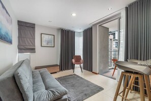 A modern living room with a grey sofa, a wooden coffee table, and a chair. There is a large window with curtains, a painting on the wall, and a view of a building outside. - Cyan 1001 - 2BR in Medell n (Medellín)