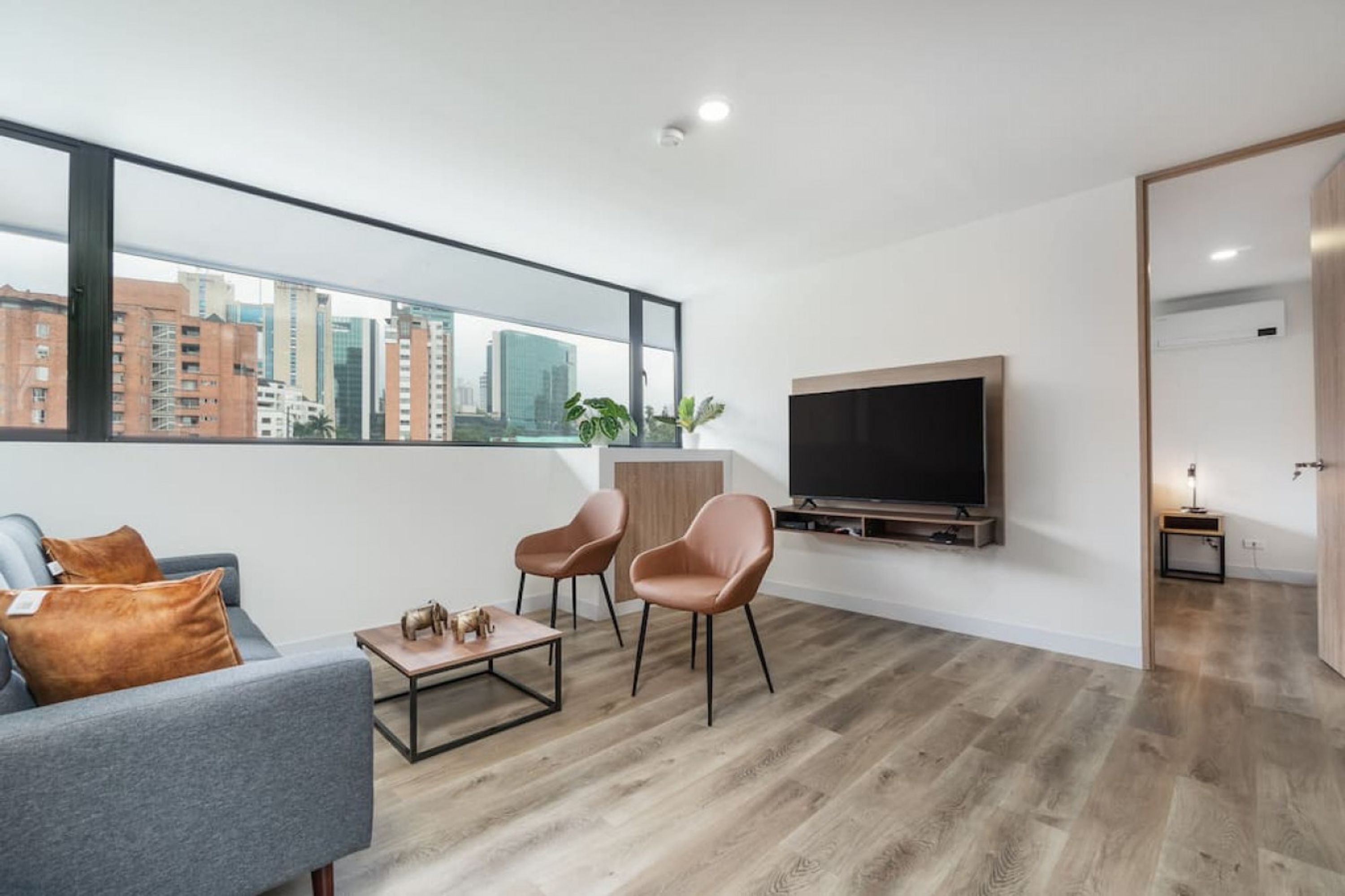 Un salon moderne doté d’une grande fenêtre, d’un téléviseur à écran plat, d’un canapé, de deux fauteuils, d’une table basse et d’un petit meuble.