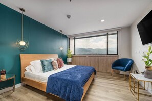 Ein modernes Schlafzimmer mit Bett, Blick auf Berge, blauem Sessel und Fernseher an der Wand.
