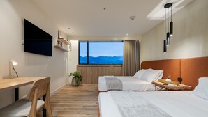 Una camera d'albergo con un letto, una scrivania, una sedia, una TV e una finestra vista montagna, più una pianta.