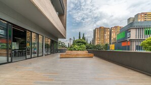 A modern building with large windows, a courtyard with a wooden bench, and high-rise buildings in the background. - Go Living Suites - 1015 (Medellín)