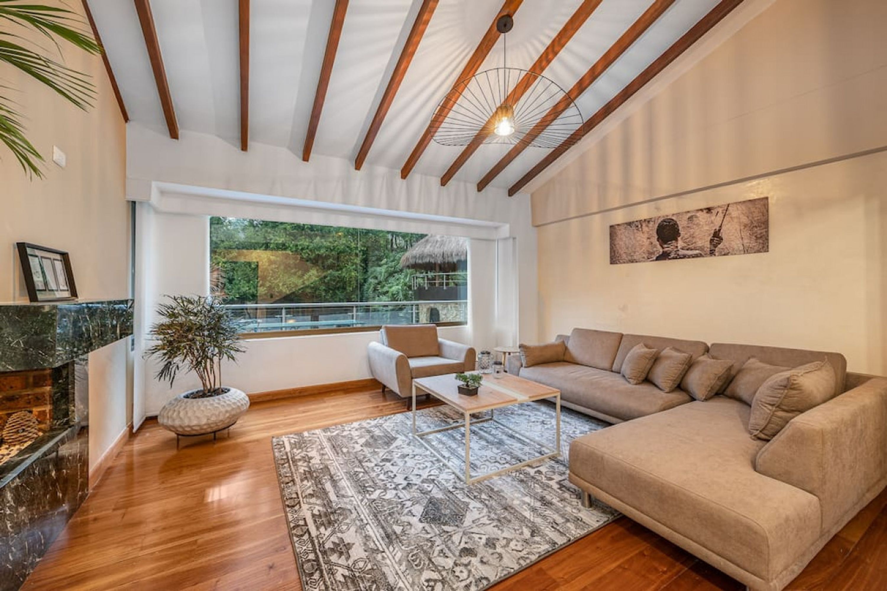 Una sala de estar moderna con techo de madera, un ventanal con vistas a la vegetación, un sofá color beige, una mesa de centro y una chimenea.