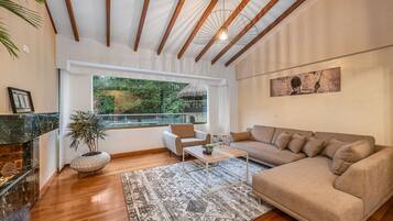 Una sala de estar moderna con techo de madera, un ventanal con vistas a la vegetación, un sofá color beige, una mesa de centro y una chimenea.