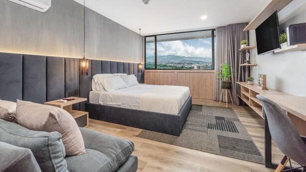Ein modernes Schlafzimmer mit Bett, Schreibtisch, Fernseher und einem großen Fenster mit Blick auf die Stadt.