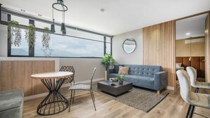 Ein modernes Wohnzimmer mit einer Couch, einem Couchtisch und Sesseln, einem Esstisch und großen Fenstern mit Pflanzen.