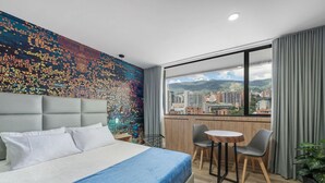 A hotel room with a bed, a small table, two chairs, a view of the city, and a colorful wall. - Go Living Suites - 1414 (Medellín)