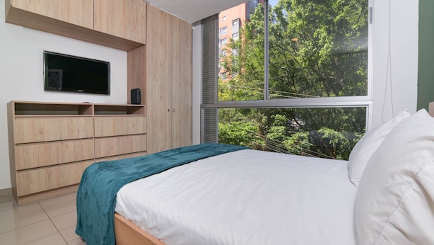 Un dormitorio con una cama, un televisor montado en la pared y una ventana con vista a árboles y edificios.
