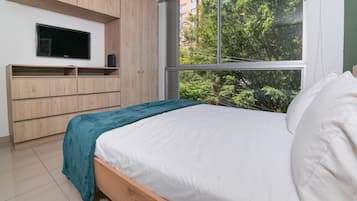 Quarto com cama, TV fixada na parede e janela com vista para árvores e edifícios.