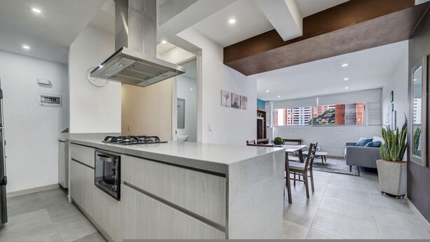 Una cocina moderna con una isla central, una zona de comedor con mesa y sillas, y un espacio de sala de estar con un sofá y un ventanal grande.