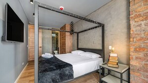 A modern bedroom with a four-poster bed, a flat-screen TV, a bedside table with a lamp, and a brick wall. - Olive Country Living - 311 (Envigado)