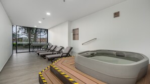 A modern indoor pool area with a hot tub, lounge chairs, and a glass wall offering a view of the outdoors. - Olive Country Living - 311 (Envigado)