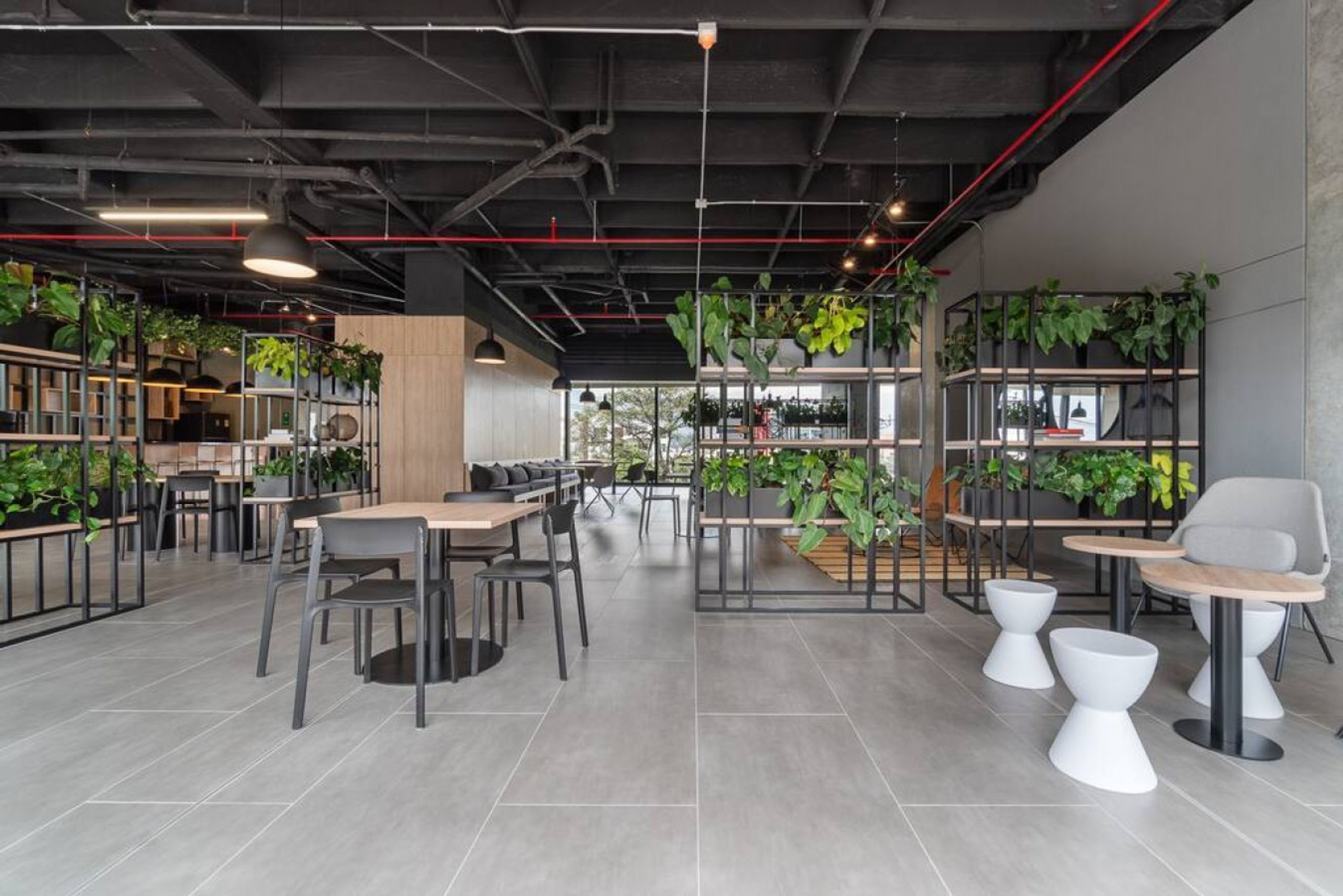Un espacio interior moderno con mesas y sillas, estanterías con plantas y un ventanal.