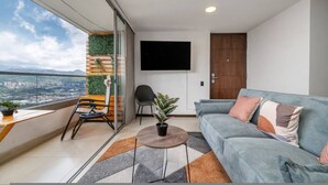 Un salon moderne avec un canapé, un téléviseur, un balcon offrant une belle vue et une petite table agrémentée d’une plante.