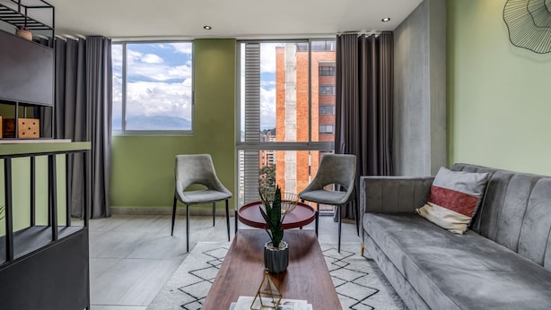Un salon moderne comprenant un canapé, deux fauteuils, une table basse et une vue sur les immeubles à travers de grandes fenêtres.