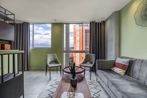 Un salon moderne comprenant un canapé, deux fauteuils, une table basse et une vue sur les immeubles à travers de grandes fenêtres.