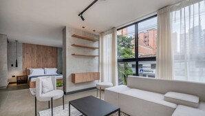 Ein modernes Wohnzimmer mit einer Couch, einem Couchtisch und einem Bett mit Holz Kopfteil. An der Wand befinden sich Holzregale und ein großes Fenster mit Vorhängen.