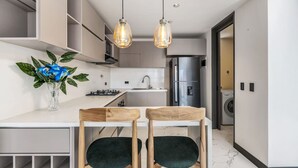 Una cocina moderna con encimera blanca, dos sillas de madera, un jarrón con flores azules y lámparas colgantes.
