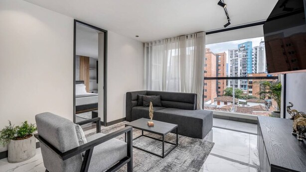 Una sala de estar moderna con sofá, sillón, mesa de centro y vistas a un paisaje urbano a través de amplios ventanales.