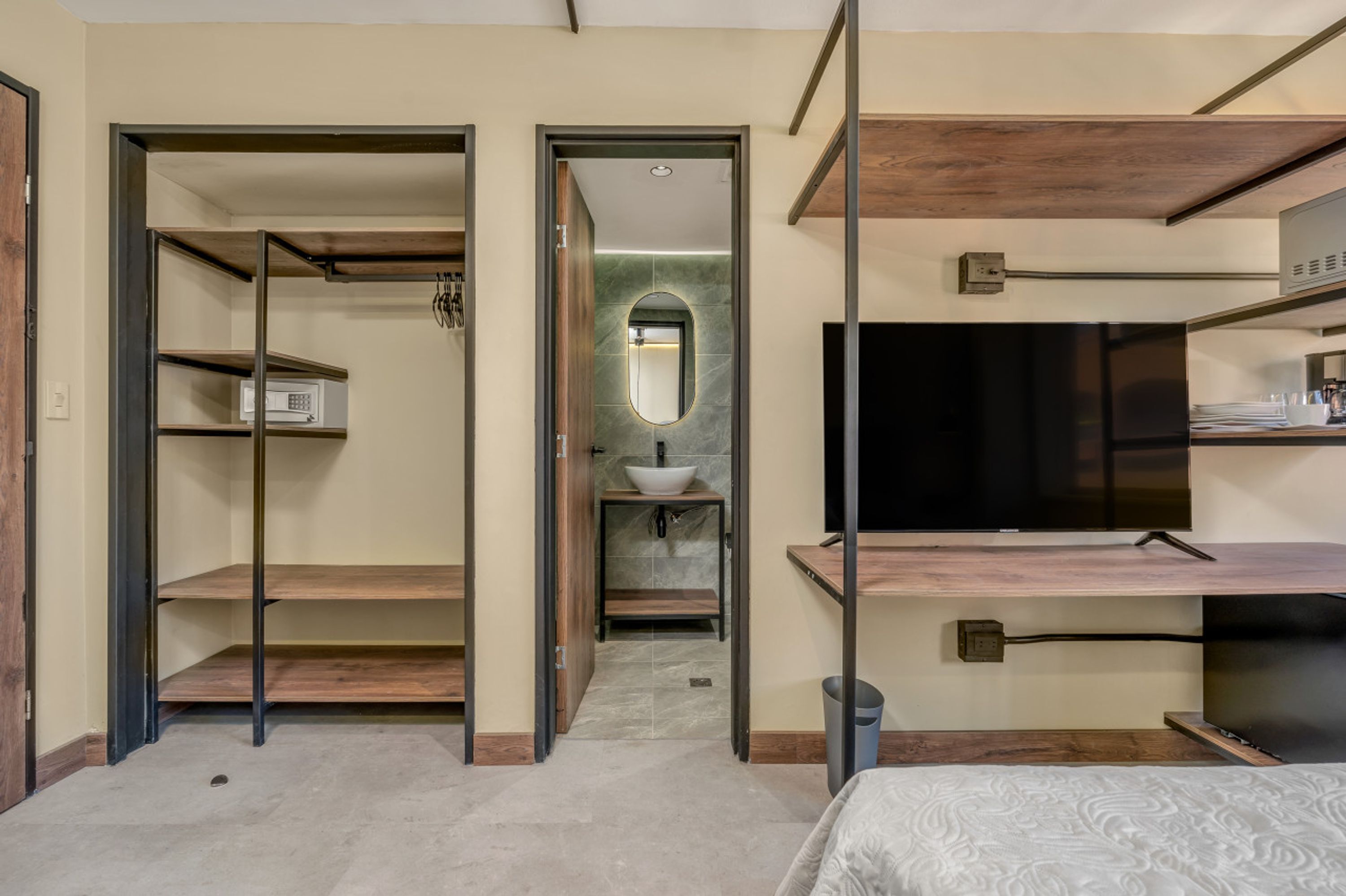 Habitación de hotel con vestidor, baño con lavamanos y espejo, y un televisor sobre un estante de madera.