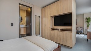 Un dormitorio moderno con cama, televisor montado en un mueble de madera, y un baño visible a través de una puerta abierta.
