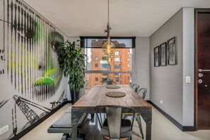 A dining area with a wooden table, metal chairs, a hanging light fixture, and a large window with a view of buildings. - Luxury 2BR Apt Near Provenza (Medellín)