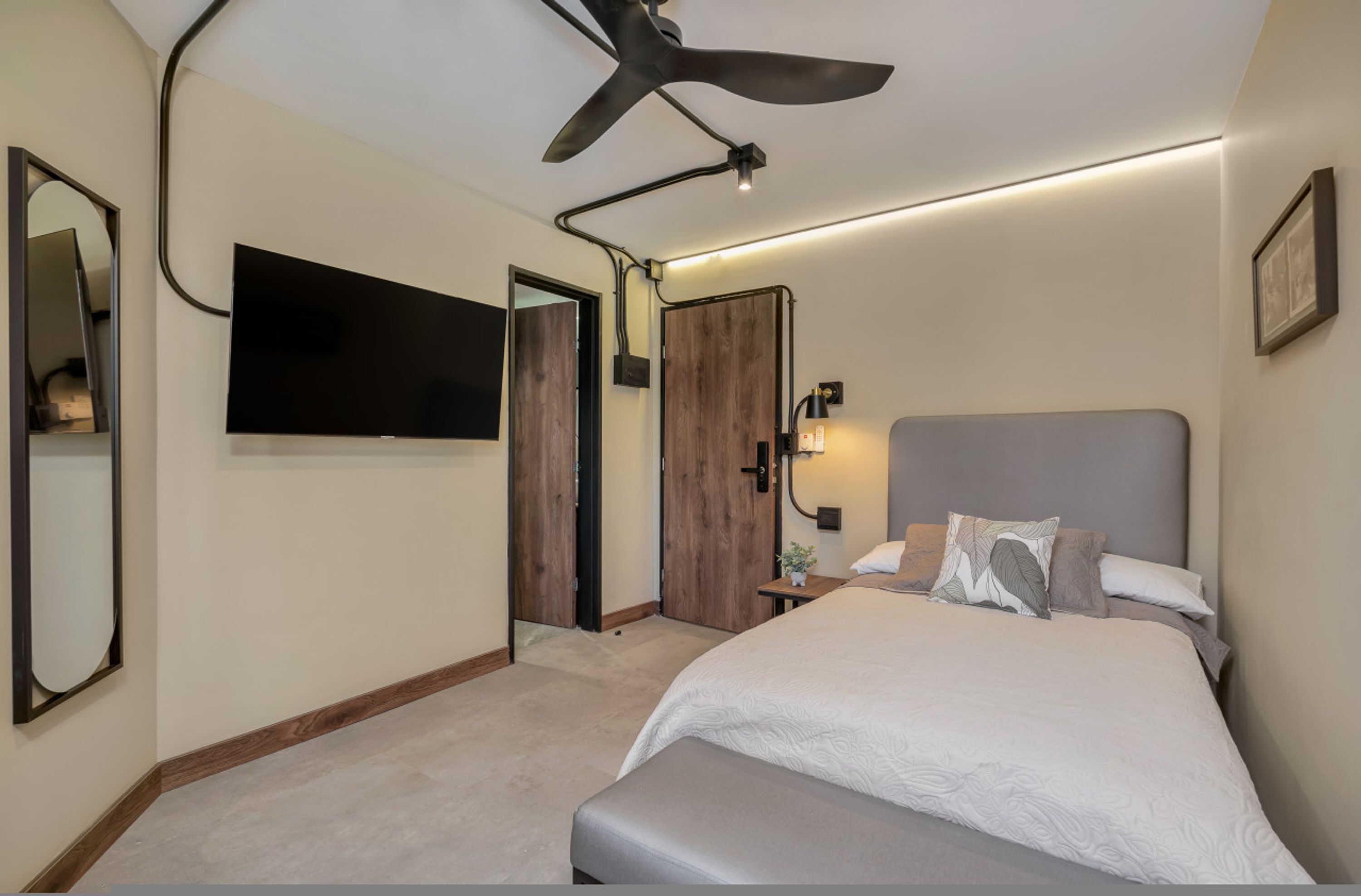Une chambre à coucher comprenant un lit, un ventilateur de plafond, un téléviseur fixé au mur, une table de chevet avec une lampe et une porte donnant sur une autre pièce.